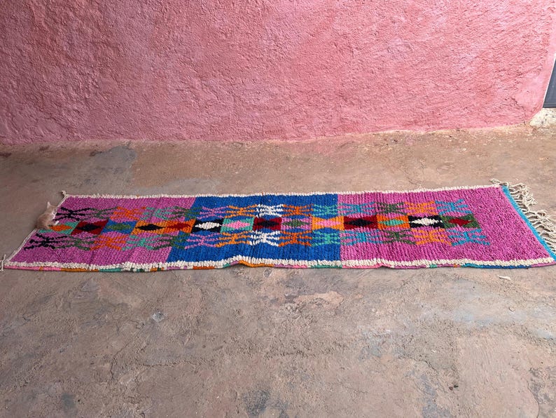 May include: A rectangular, hand-woven rug with a vibrant geometric pattern. The rug features a central blue section flanked by pink borders. The design incorporates orange, green, and white accents, with fringed edges.