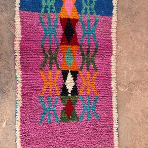 May include: A rectangular, hand-woven rug with a vibrant geometric design. The rug features a central pattern of diamonds and crosses in shades of pink, blue, orange, green, and black. The edges are trimmed with a white fringe.