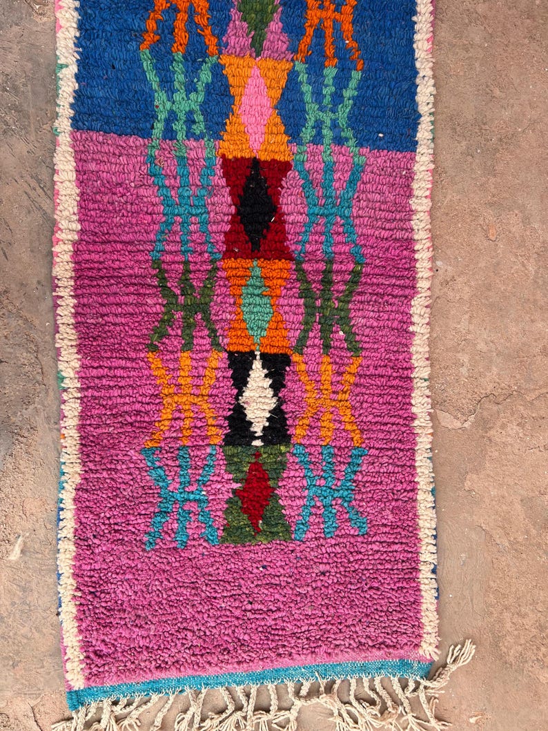 May include: A rectangular, hand-woven rug with a vibrant geometric design. The rug features a central pattern of diamonds and crosses in shades of pink, blue, orange, green, and black. The edges are trimmed with a white fringe.