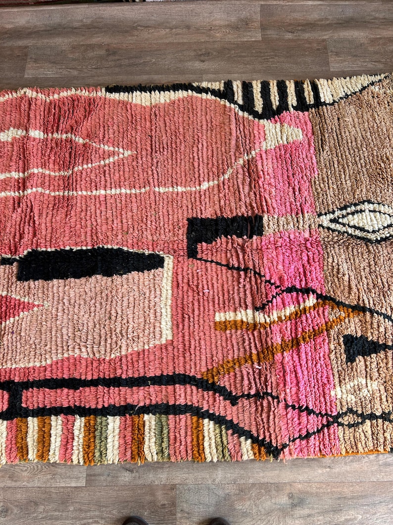 May include: A colorful, abstract rug with geometric shapes in shades of pink, brown, black, and white. The rug has a hand-woven texture and a modern design.