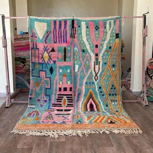 Op de afbeelding: Een rechthoekig vloerkleed met een levendige turquoise achtergrond en een complex geometrisch patroon in roze, oranje, geel, zwart en wit. Het ontwerp bevat diverse abstracte vormen, lijnen en symbolen, afgewerkt met geknoopte franjes.