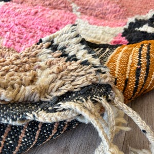 May include: Close-up of the edge of a colorful woven rug with pink, white, brown, orange, and black stripes. The rug is folded and the fringe is visible.