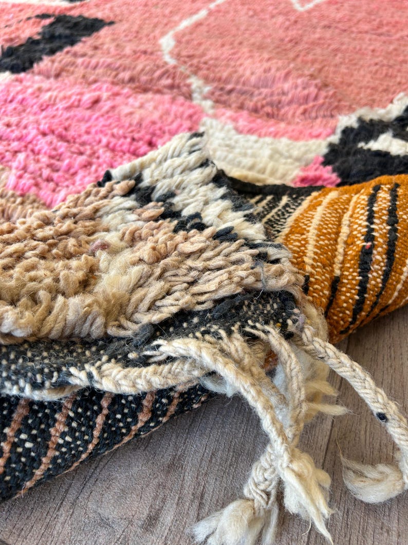 May include: Close-up of the edge of a colorful woven rug with pink, white, brown, orange, and black stripes. The rug is folded and the fringe is visible.