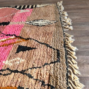 May include: A hand-woven Moroccan rug with a geometric pattern in shades of pink, brown, black, and white. The rug has a fringe border.