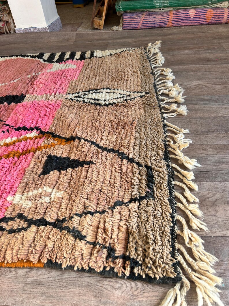 May include: A hand-woven Moroccan rug with a geometric pattern in shades of pink, brown, black, and white. The rug has a fringe border.