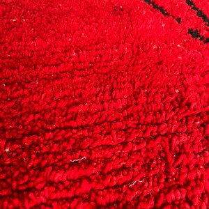 Peut inclure: Une vue rapproch&eacute;e d'un tapis rouge vif avec une texture &agrave; poils touffet&eacute;s &eacute;pais. Une partie d'un motif graphique est visible en haut, pr&eacute;sentant une forme gris clair soulign&eacute;e par un trait noir &eacute;pais sur le fond rouge vif du tapis.