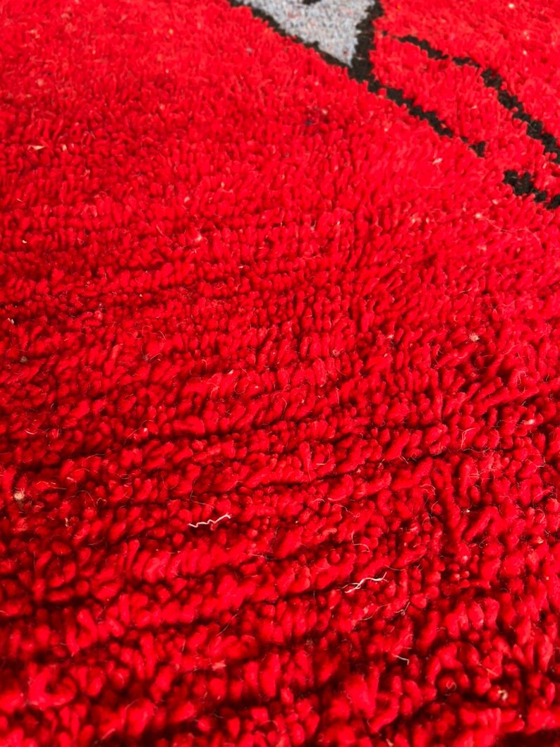 Peut inclure: Une vue rapproch&eacute;e d'un tapis rouge vif avec une texture &agrave; poils touffet&eacute;s &eacute;pais. Une partie d'un motif graphique est visible en haut, pr&eacute;sentant une forme gris clair soulign&eacute;e par un trait noir &eacute;pais sur le fond rouge vif du tapis.