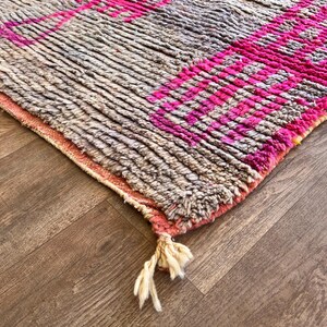 May include: A close-up of a textured rug with a geometric pattern. The rug features a combination of grey and pink hues, with the pink forming bold stripes and shapes. The rug has a fringe detail at the edge.