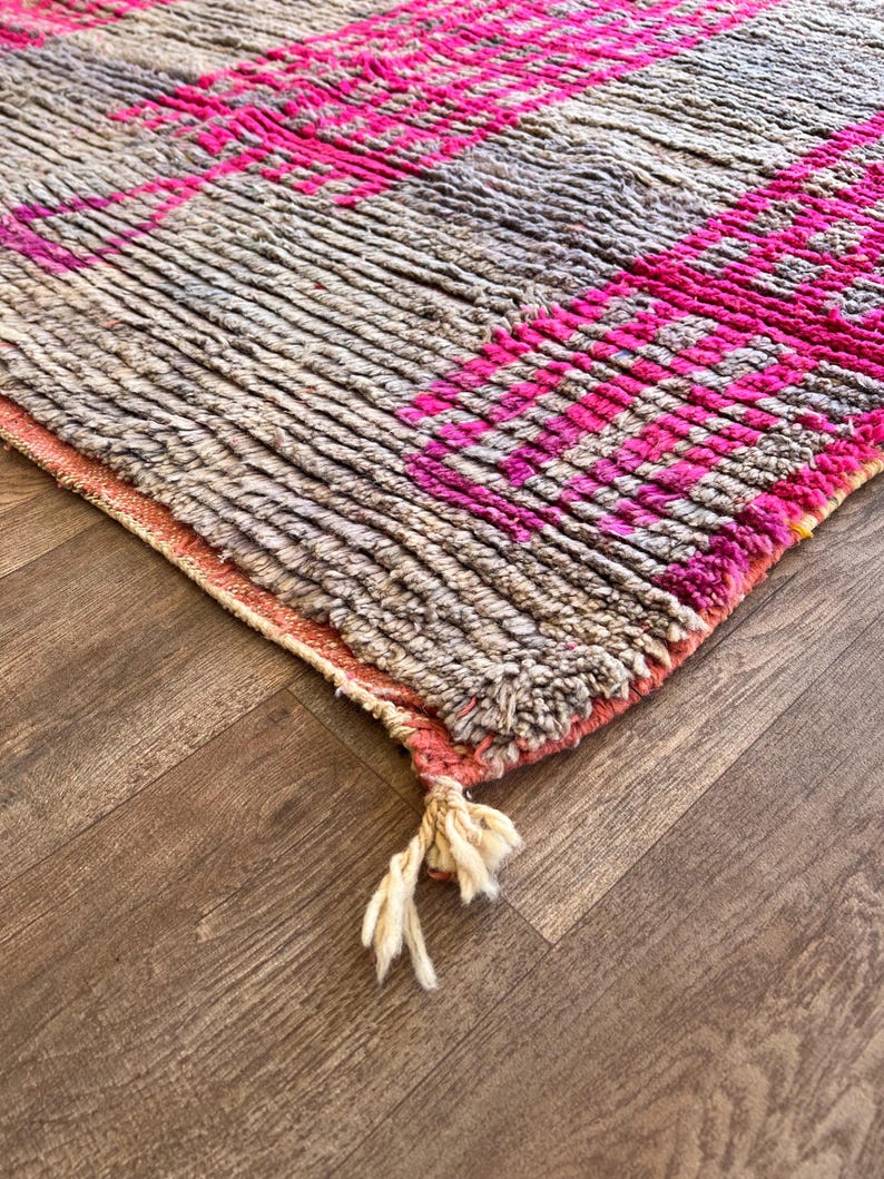 May include: A close-up of a textured rug with a geometric pattern. The rug features a combination of grey and pink hues, with the pink forming bold stripes and shapes. The rug has a fringe detail at the edge.