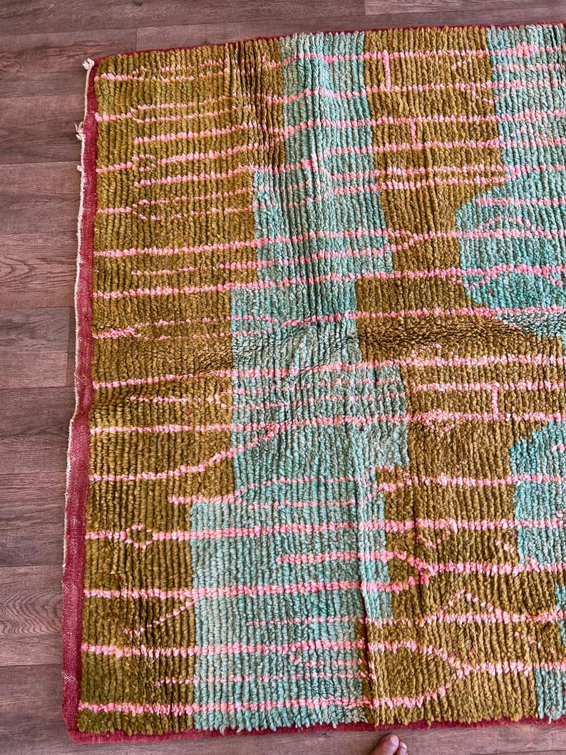 May include: A rectangular rug with a textured, woven design. The rug features a colour block pattern with sections of olive green, light blue, and pink stripes. The edges are trimmed with a red border.