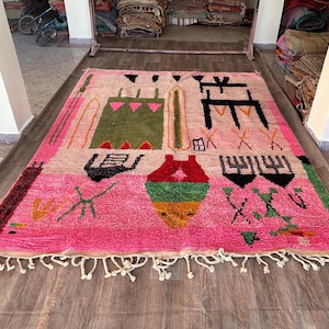 May include: A rectangular area rug featuring a vibrant pink background with various abstract geometric shapes and symbols in green, black, red, yellow, and orange. The rug has a thick pile texture and includes white fringe along the bottom edge.
