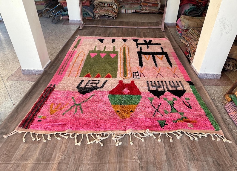 May include: A rectangular area rug featuring a vibrant pink background with various abstract geometric shapes and symbols in green, black, red, yellow, and orange. The rug has a thick pile texture and includes white fringe along the bottom edge.