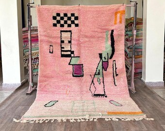 May include: A pink Moroccan rug with a geometric design in black, green, and purple. The rug has a fringe border and is made of wool.