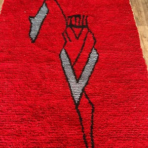 Peut inclure: Un tapis rectangulaire &agrave; poils longs d'un rouge profond. Le tapis pr&eacute;sente un grand motif graphique abstrait en noir et gris clair, repr&eacute;sentant une silhouette stylis&eacute;e avec une t&ecirc;te pointue et des lignes angulaires sur le fond rouge vif.