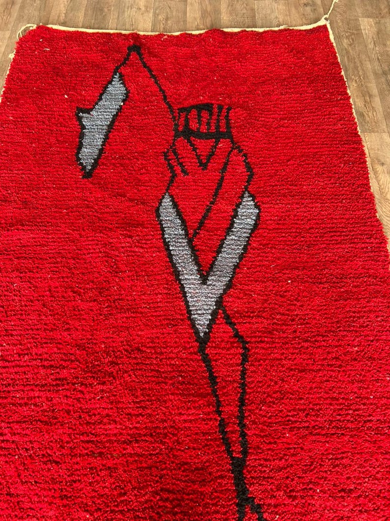 Peut inclure: Un tapis rectangulaire &agrave; poils longs d'un rouge profond. Le tapis pr&eacute;sente un grand motif graphique abstrait en noir et gris clair, repr&eacute;sentant une silhouette stylis&eacute;e avec une t&ecirc;te pointue et des lignes angulaires sur le fond rouge vif.