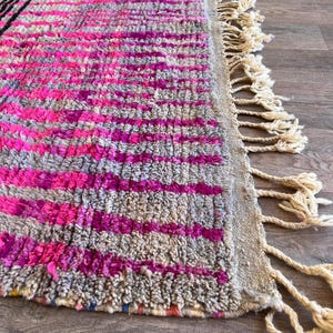 May include: Close-up of a colourful rug with a geometric design. The rug has a textured surface with pink, grey, and black stripes. Cream-coloured tassels form a fringe along one edge. The rug's design adds a vibrant touch.