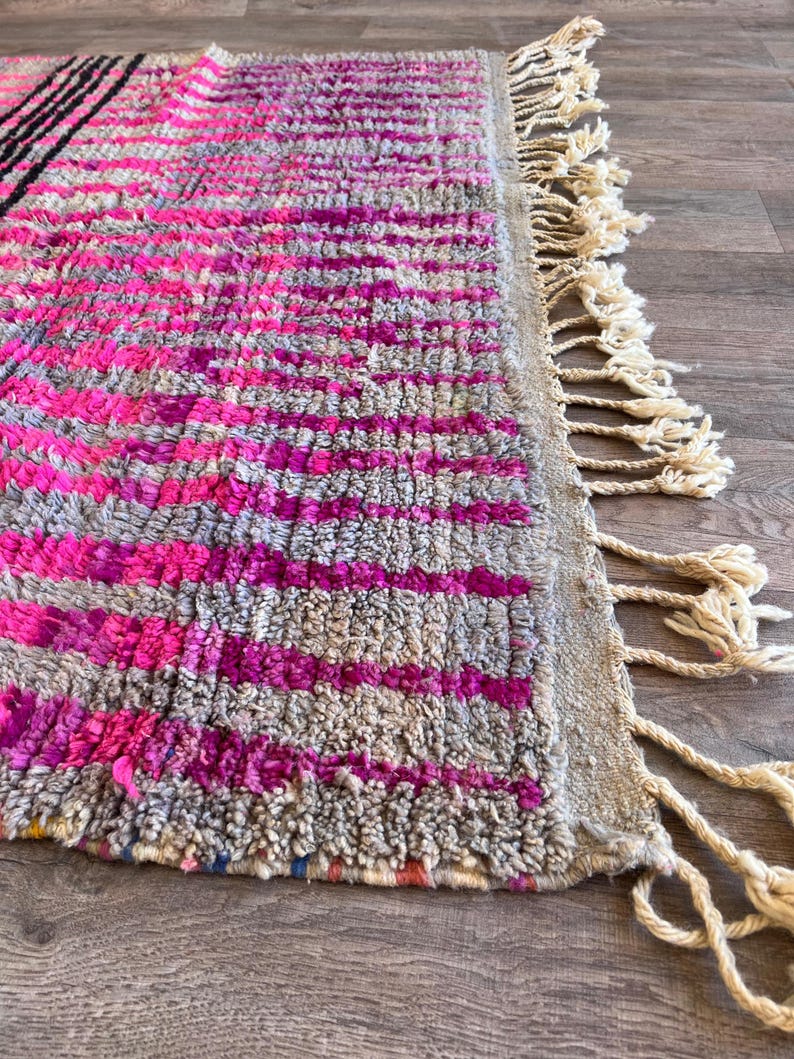 May include: Close-up of a colourful rug with a geometric design. The rug has a textured surface with pink, grey, and black stripes. Cream-coloured tassels form a fringe along one edge. The rug's design adds a vibrant touch.