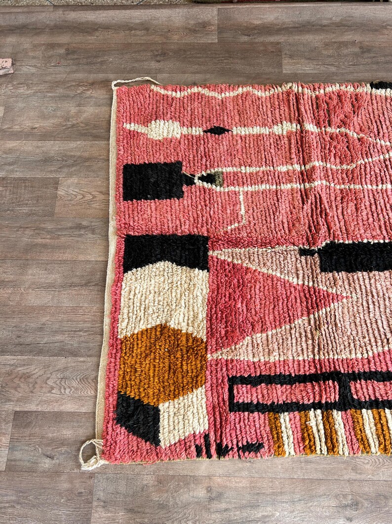 May include: A pink, black, white, and brown geometric rug with a textured, woven design. The rug has a fringe border.