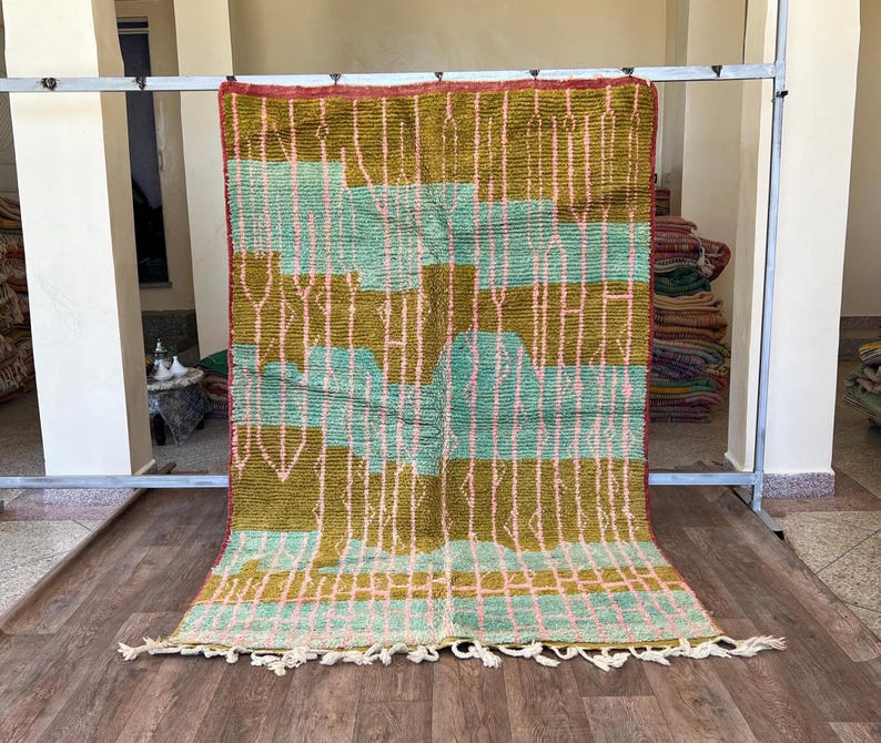May include: A rectangular, hand-woven rug with a geometric pattern. The rug features a combination of olive green, light blue, and pink colours. The design includes vertical and horizontal lines, with a fringe along the bottom edge. The rug is displayed on a wooden floor.
