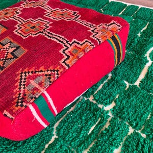 May include: A red square floor cushion with a geometric pattern in orange, black, and white. The cushion is placed on a green rug with a white grid pattern. The cushion has a red side with a striped accent.