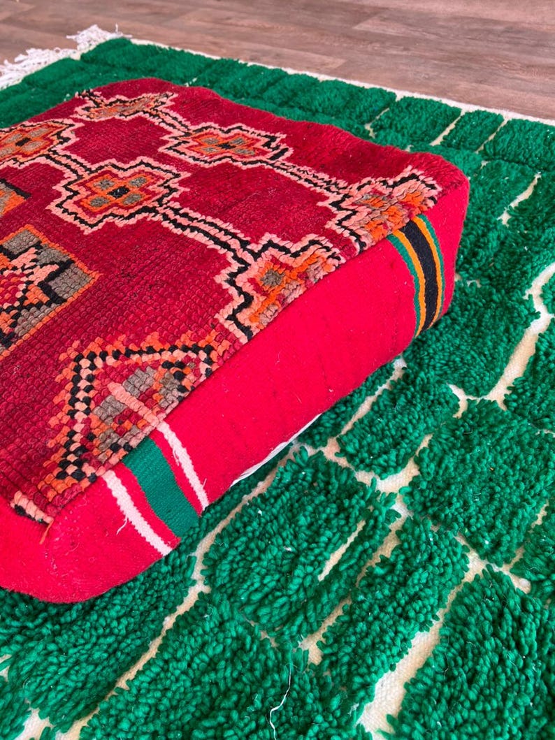 May include: A red square floor cushion with a geometric pattern in orange, black, and white. The cushion is placed on a green rug with a white grid pattern. The cushion has a red side with a striped accent.