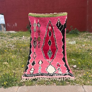 Puede incluir: Una vibrante alfombra tejida a mano con un dise&ntilde;o central rosa y rojo, acentuado por patrones geom&eacute;tricos en blanco y negro. La alfombra tiene un borde verde y un borde con flecos. Se exhibe al aire libre sobre hierba.