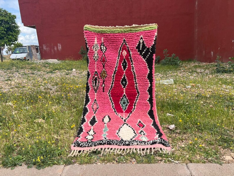 Puede incluir: Una vibrante alfombra tejida a mano con un dise&ntilde;o central rosa y rojo, acentuado por patrones geom&eacute;tricos en blanco y negro. La alfombra tiene un borde verde y un borde con flecos. Se exhibe al aire libre sobre hierba.