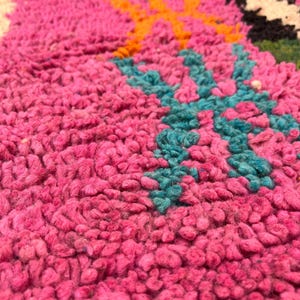 May include: Close-up of a vibrant pink rug with a textured, plush surface. The rug features a colourful geometric design with teal, orange, green, and black accents. The rug's design is a mix of floral and geometric patterns.