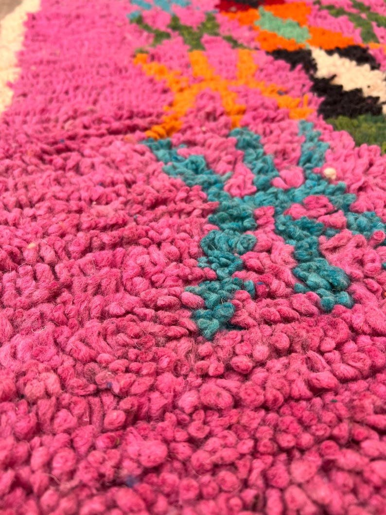 May include: Close-up of a vibrant pink rug with a textured, plush surface. The rug features a colourful geometric design with teal, orange, green, and black accents. The rug's design is a mix of floral and geometric patterns.