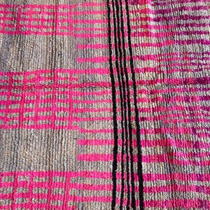 May include: A rectangular rug with a textured surface. The rug features a pattern of pink and grey rectangles, with a vertical stripe of black lines on the right side. The rug is made of a woven material.