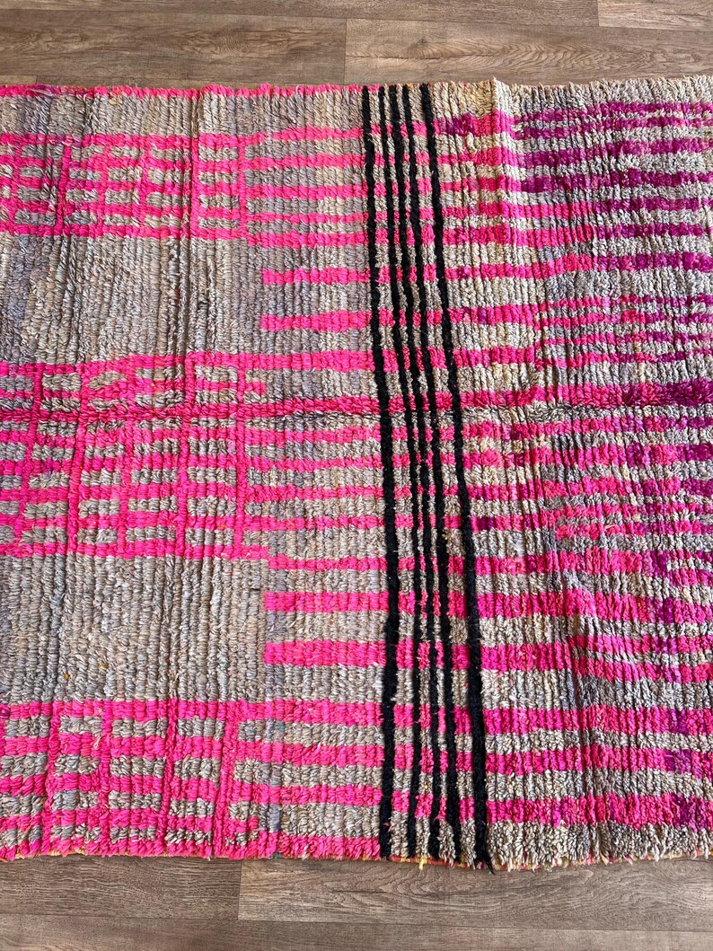 May include: A rectangular rug with a textured surface. The rug features a pattern of pink and grey rectangles, with a vertical stripe of black lines on the right side. The rug is made of a woven material.