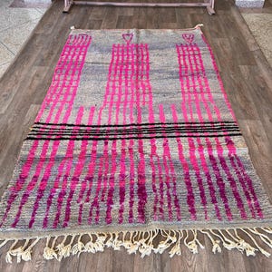 May include: A rectangular grey rug with a pink and black geometric pattern. The rug features vertical pink stripes and three black horizontal stripes. The rug has a fringe along the bottom edge.