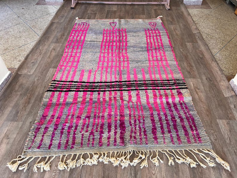 May include: A rectangular grey rug with a pink and black geometric pattern. The rug features vertical pink stripes and three black horizontal stripes. The rug has a fringe along the bottom edge.
