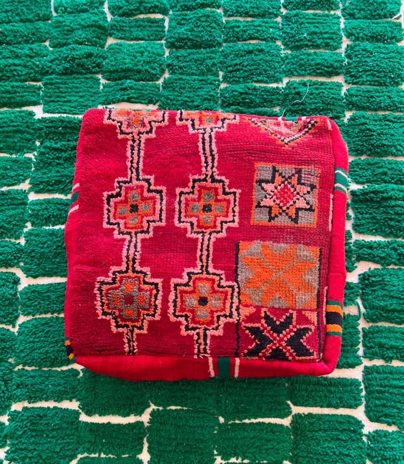 May include: A square, red decorative pillow with a geometric pattern in orange, gray, and black. The pillow is placed on a green, textured rug. The pillow's design features repeating cross-like shapes and starburst motifs.