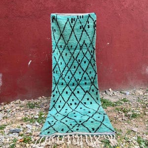 Peut inclure: Tapis rectangulaire turquoise avec un motif de losanges noirs et des franges. Le tapis est fait d'une matière épaisse et texturée et est présenté contre un mur rouge et une surface de gravier.
