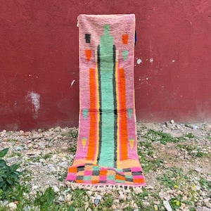 Peut inclure: Un tapis rectangulaire coloré avec un fond rose et un motif vertical central. Le motif présente des rayures orange, vertes et jaunes, avec des formes géométriques noires et roses. Le tapis a une bordure frangée et est présenté contre un mur rouge.