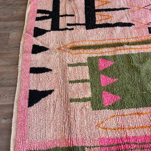 May include: A hand-knotted wool rug featuring a light pink base with a high-pile texture. The design includes black geometric shapes, a large olive green rectangular block with three pink triangles, and orange linear accents along the edges.