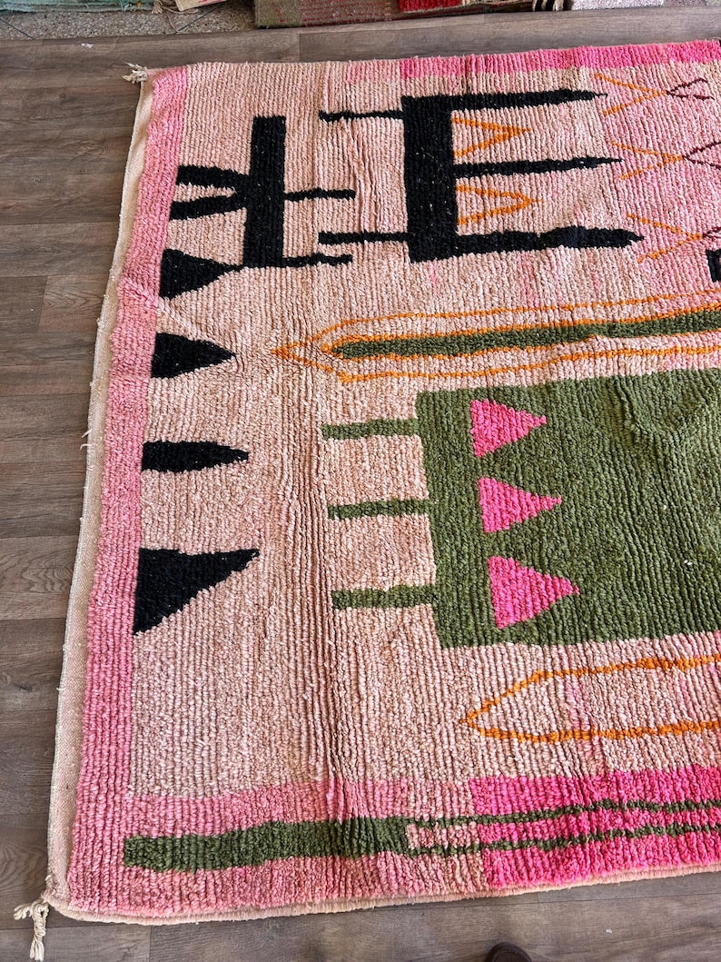May include: A hand-knotted wool rug featuring a light pink base with a high-pile texture. The design includes black geometric shapes, a large olive green rectangular block with three pink triangles, and orange linear accents along the edges.