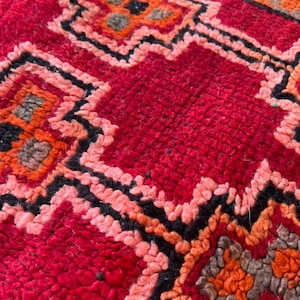 May include: Close-up of a vibrant red rug with a geometric pattern. The design features repeating shapes in shades of orange, black, and pink. The rug has a textured, woven appearance, suggesting a handmade or artisanal quality.