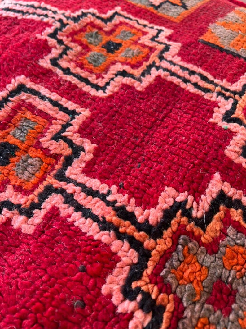 May include: Close-up of a vibrant red rug with a geometric pattern. The design features repeating shapes in shades of orange, black, and pink. The rug has a textured, woven appearance, suggesting a handmade or artisanal quality.