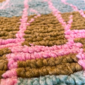 May include: Close-up of a colourful rug with a geometric pattern. The rug features a combination of pink, brown, and teal colours. The texture of the rug appears soft and plush, with visible yarn strands creating a textured surface.
