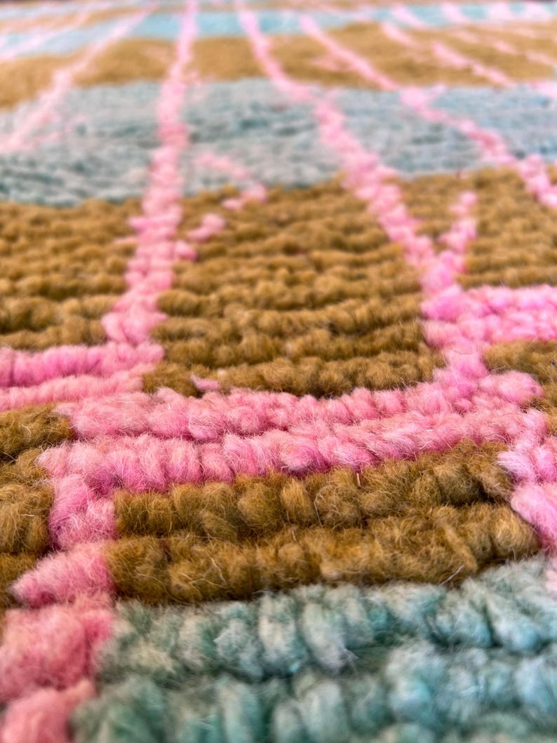May include: Close-up of a colourful rug with a geometric pattern. The rug features a combination of pink, brown, and teal colours. The texture of the rug appears soft and plush, with visible yarn strands creating a textured surface.