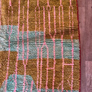 May include: Close-up of a textured rug with a geometric pattern. The rug features vertical stripes in pink and olive green, with sections of turquoise blue. The edges are trimmed with a red border, creating a vibrant and colorful design.