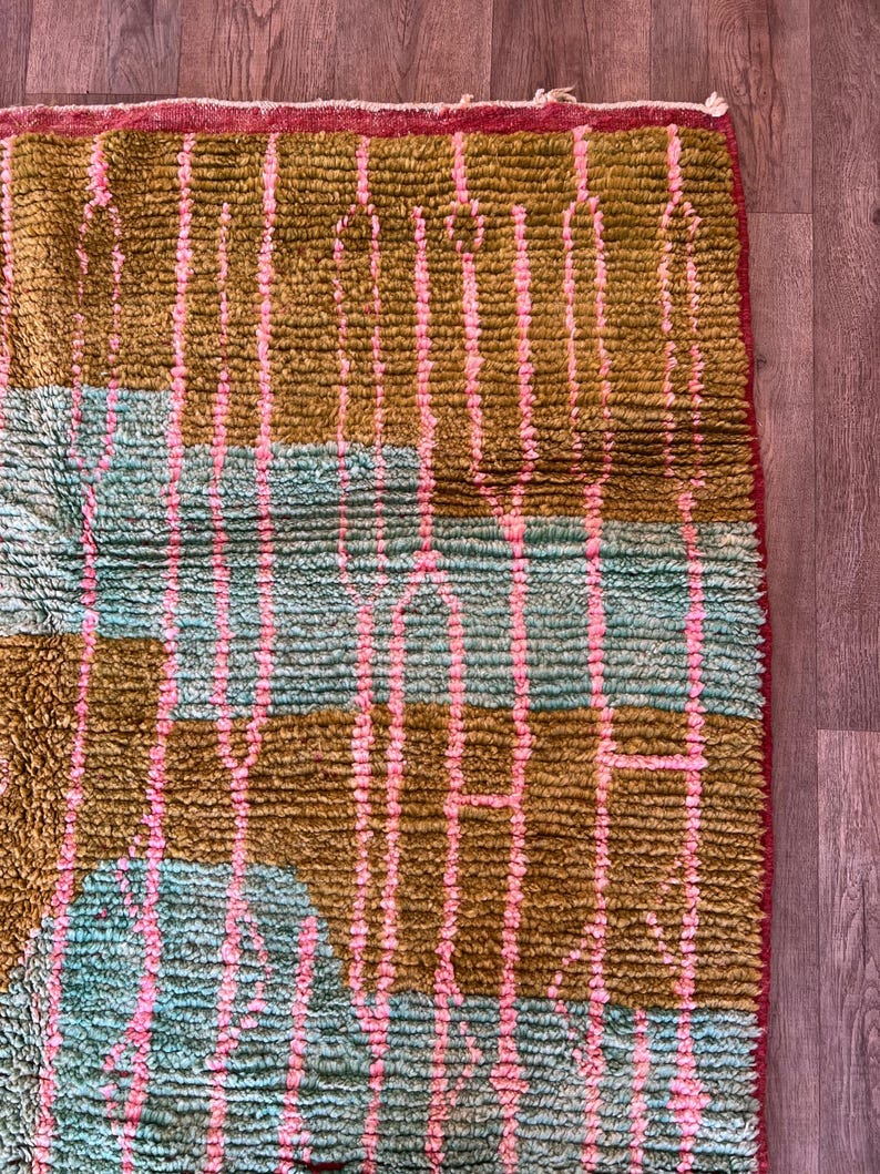 May include: Close-up of a textured rug with a geometric pattern. The rug features vertical stripes in pink and olive green, with sections of turquoise blue. The edges are trimmed with a red border, creating a vibrant and colorful design.