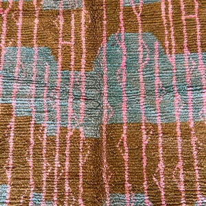 May include: A close-up of a textured rug with a repeating pattern. The rug features a teal background with brown rectangular shapes and vertical pink lines. The rug has a soft, plush appearance.