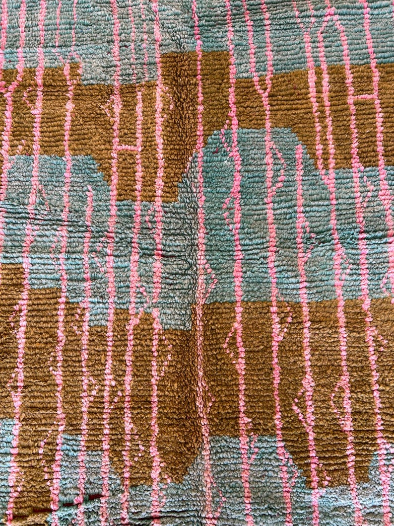 May include: A close-up of a textured rug with a repeating pattern. The rug features a teal background with brown rectangular shapes and vertical pink lines. The rug has a soft, plush appearance.