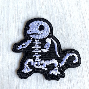 Puede incluir: Parche bordado negro con un diseño de esqueleto en morado claro y blanco. El parche tiene la forma de un personaje de dibujos animados. Tiene un borde negro y es probablemente para ropa o accesorios.