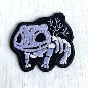 Puede incluir: Un parche bordado negro con un esqueleto blanco de una criatura de dibujos animados. La criatura está de pie sobre sus patas traseras con los brazos levantados. El esqueleto está delineado en blanco y tiene un brillo púrpura.