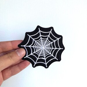 May include: A black and white embroidered spiderweb patch. The spiderweb is white and the background is black.