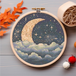Pode incluir: Um aro de bordado de ponto cruz com uma lua crescente e estrelas sobre um fundo azul escuro. A lua é de cor amarelo claro e as estrelas são brancas. O fundo está cheio de nuvens brancas.